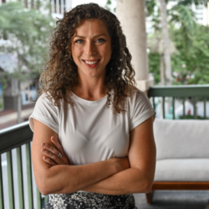 A woman with curly hair stands on a balcony with her arms crossed, wearing a white t-shirt and patterned skirt. Trees and buildings are visible in the background.