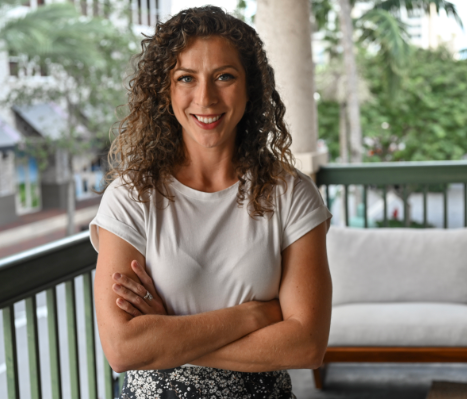 A woman with curly hair stands on a balcony with her arms crossed, wearing a white t-shirt and patterned skirt. Trees and buildings are visible in the background.