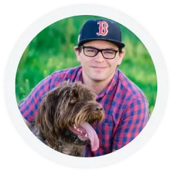 A man in glasses and a Boston Red Sox cap sits outdoors with a brown, long-haired dog.