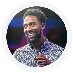 A man with a beard and short curly hair smiles and gestures while wearing a patterned shirt; the background is blurred.