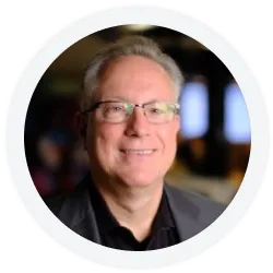 Smiling middle-aged man with glasses and gray hair, wearing a dark jacket and shirt, posed indoors with a blurred background.