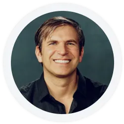 A man with short brown hair wearing a black shirt smiles at the camera against a dark green background.