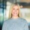 A woman with straight blonde hair wearing a grey sweater smiles at the camera, standing indoors with a blurred background.