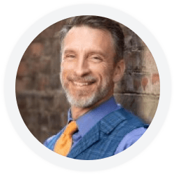 A man with short brown hair and a gray beard, wearing a blue vest, lavender shirt, and orange tie, smiling and standing against a brick wall.