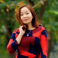 Woman with shoulder-length brown hair wearing a red and navy patterned sweater stands outdoors, touching her hair, with trees in the background.