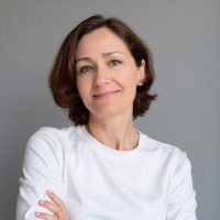 Woman with short brown hair wearing a white shirt stands in front of a plain gray background, looking at the camera with a slight smile and arms crossed.