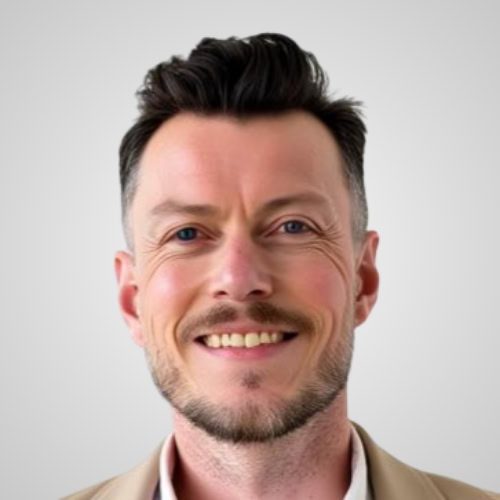 A man with short dark hair and a trimmed beard smiles at the camera, wearing a beige jacket and standing in front of a plain light gray background.