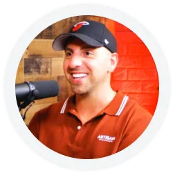 A man wearing a black cap and red collared shirt speaks into a microphone, sitting in front of a brick wall and wooden panel background.