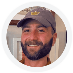 Man with a beard and mustache, wearing a gray baseball cap and brown shirt, smiling at the camera with framed pictures in the background.